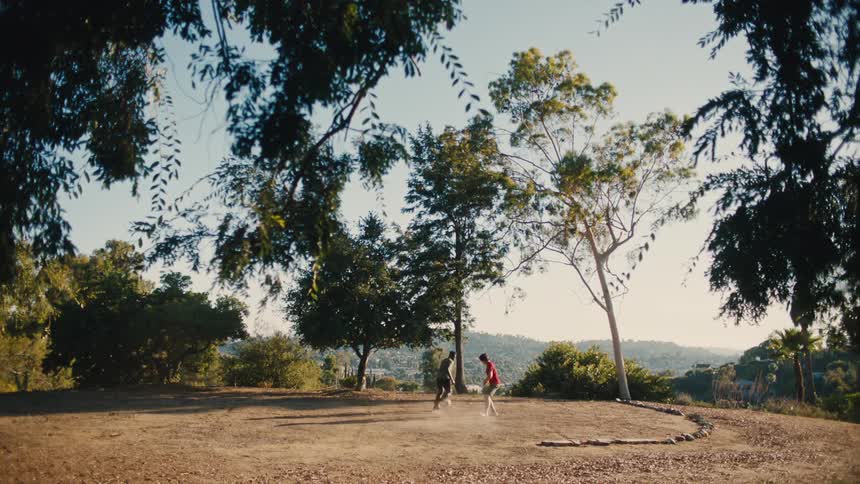 Two youths play one on one soccer on a dusty field overlooking a valley