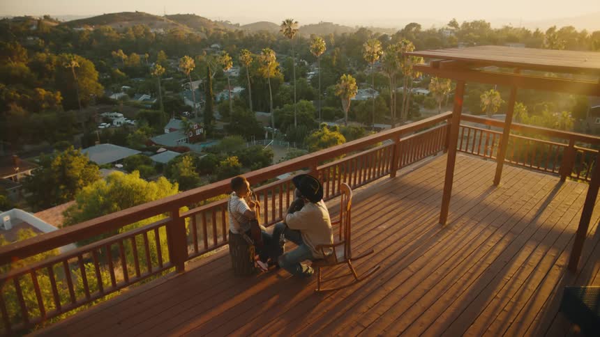 A father holding a guitar sitting in a rocking chair on a large porch teaching his son how to play guitar as the sun sets