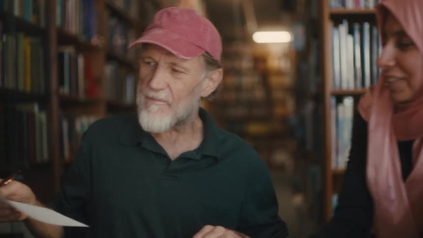 A woman with a pink turban and a man with a pink cap who are sitting in a library talking and are happy