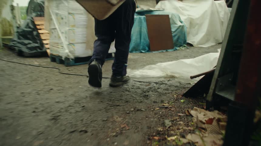 A groundlevel view of a persons lower legs and feet wearing muddy boots carrying a mattress through a yard