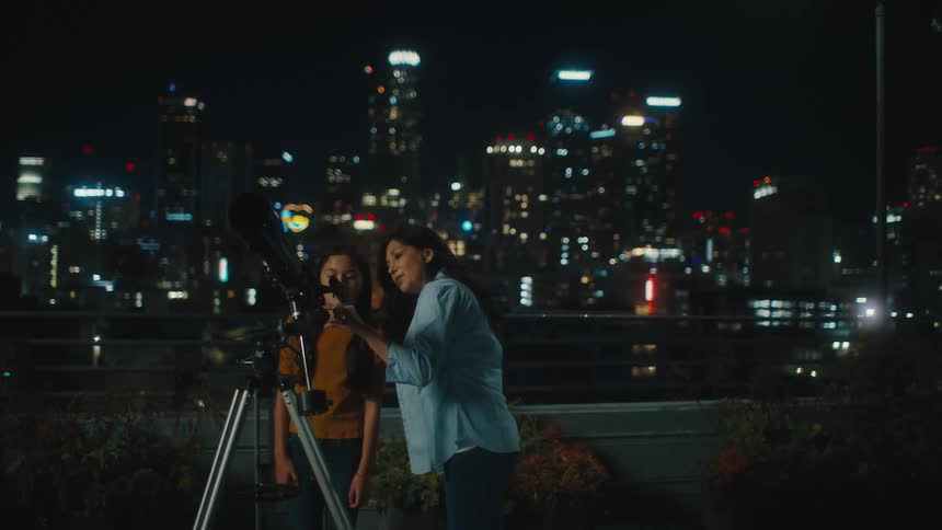 A mother teaching her daughter how to use a telescope with city lights in the background at night