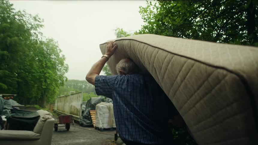 Elderly man carrying a mattress through a yard