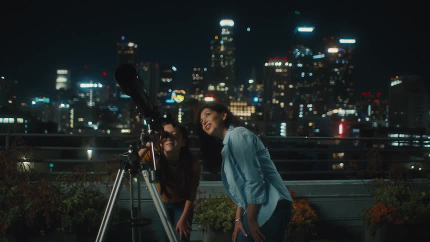 Woman next to a girl who is looking into the telescope at night in a city with skyscrapers in the background