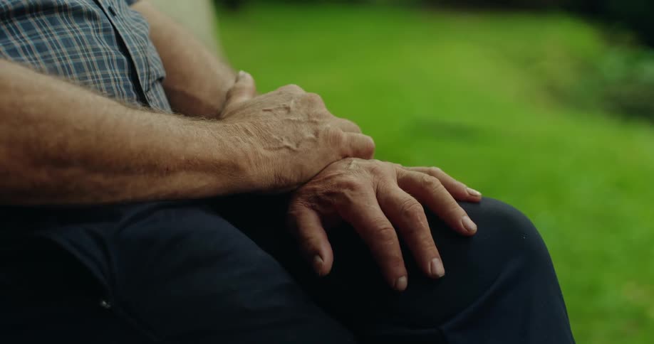 Close up of an older mans hands placed on his lap as he sits outside