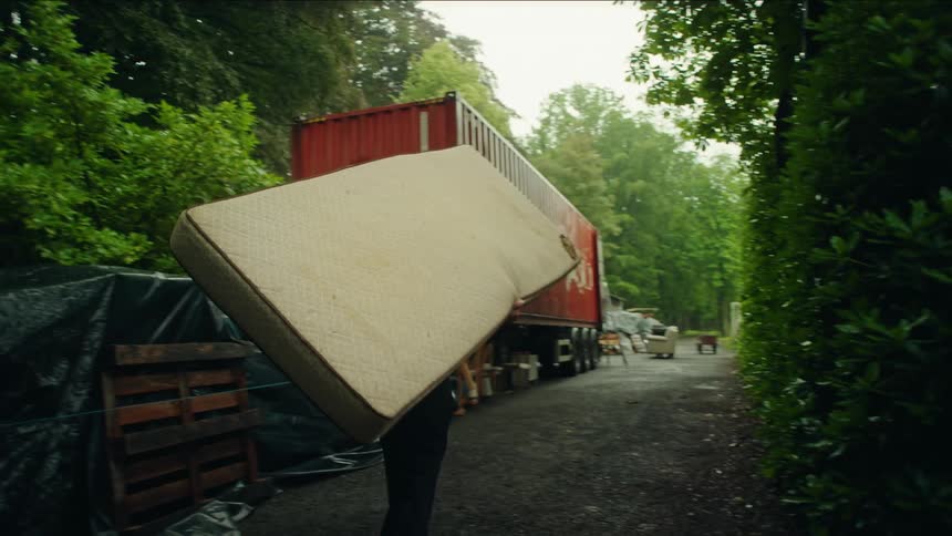 A man carrying a white mattress down a dirt path with vegetation on either side and toward a red shipping container