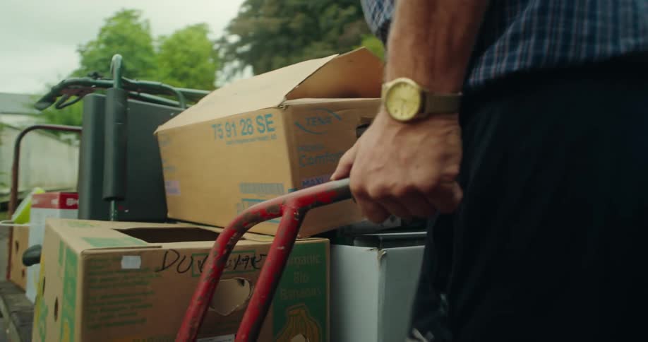 An elderly man wheels a cart stacked with boxes through a yard