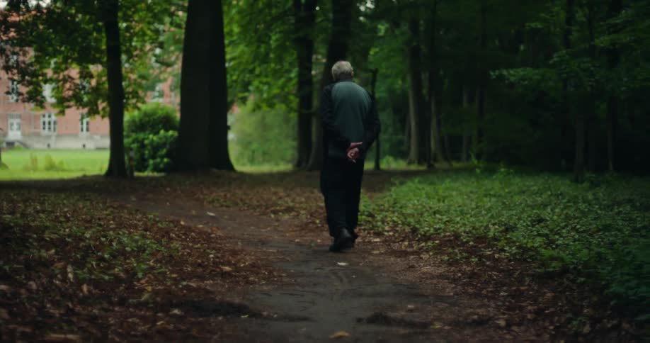 An elderly man with his hands clasped behind his back is walking on a path between some trees