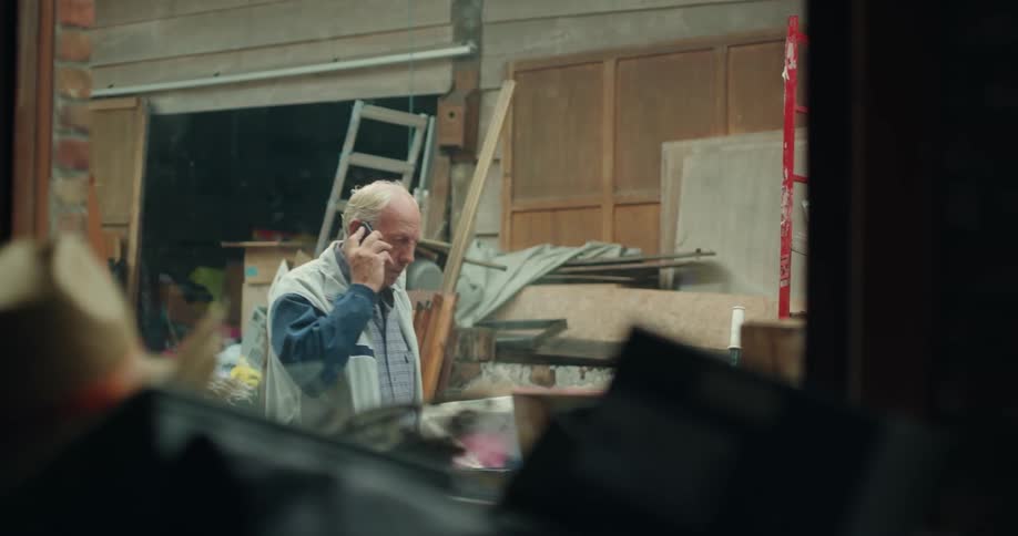 An elderly man is walking past a shed with a lot of junk around him while holding a cell phone to his ear