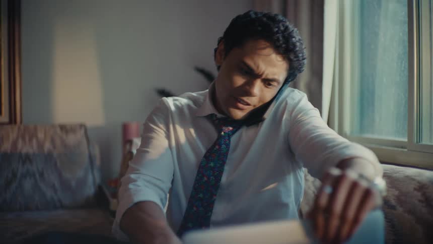 A hispanic man talks on his smartphone while sitting on his couch and typing on a laptop