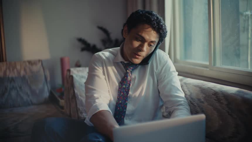 A man in a shirt and tie talking on his cellphone walking through a living room to sit on a couch and type on his laptop before getting up