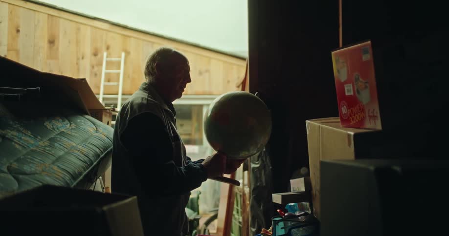 An elderly man is in a shed and he is holding a globe and placing it on top of a box in front of him