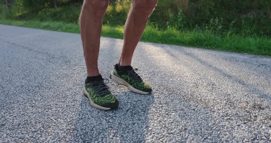 A mans legs and feet in green sneakers pacing in place on a roadway as the sun shines through the trees