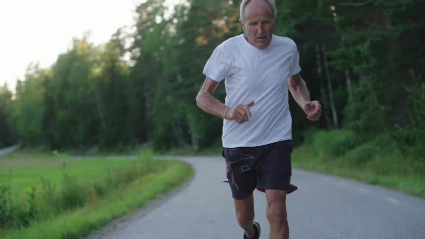 Older man runs along a highway passing between a field and a grove of trees