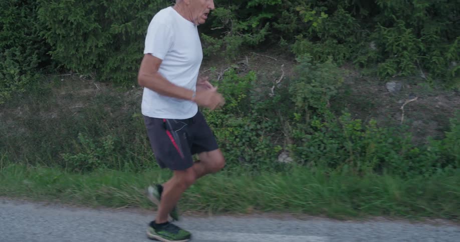 An elderly man in a white shirt and black shorts is jogging along a roadside