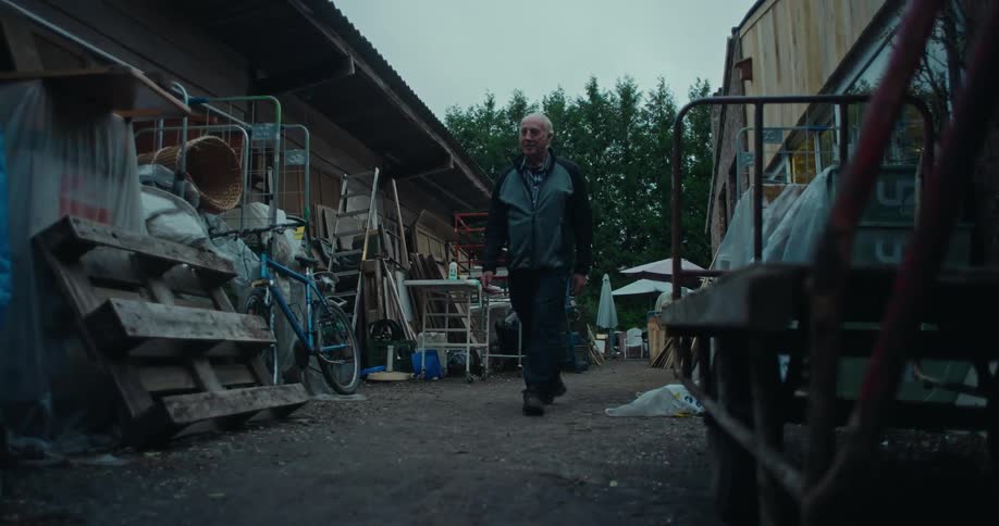 An older man walking on a dirt path between pallets and other items are stacked between buildings