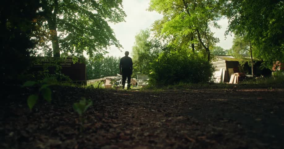 An elderly man walks down a rural dirt road on a summer day with a yard in the background