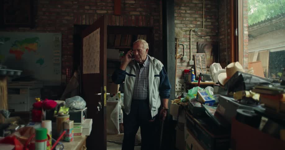 An elderly man standing in a cluttered room talking on the phone