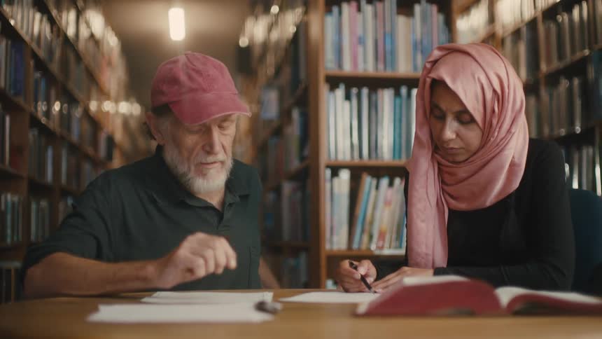 A bearded man explains a document to a woman wearing a pink headscarf in a library