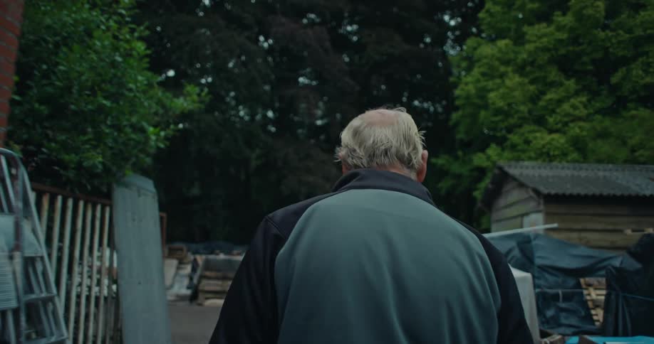The backside of a man as he walks next to a brick building through piles of stuff in an outdoor area