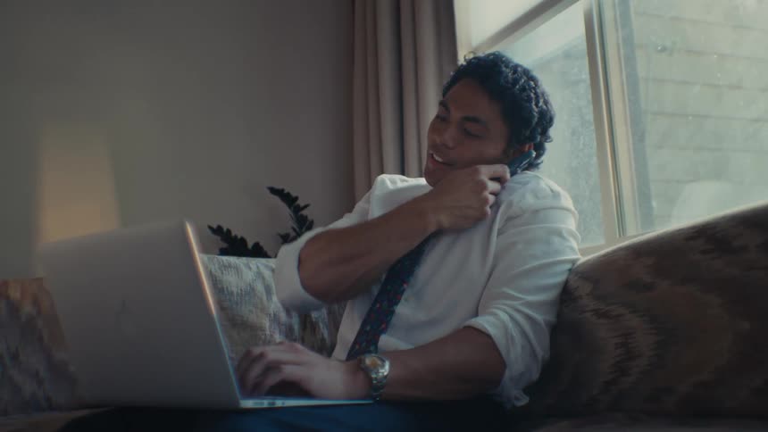 A man sitting on his couch while talking on his cellphone and typing on his laptop