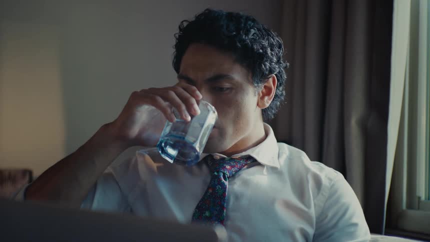 A man in a dress shirt and tie sitting on his couch drinking from a glass while looking at his laptop furrowing his brow