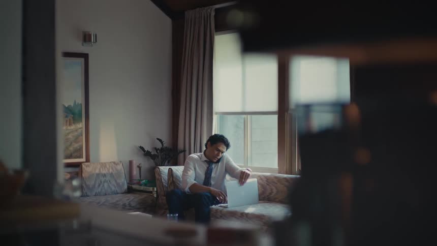 A man talking on his cellphone while walking through the living room to sit on the couch and type on his laptop