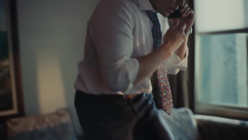 A young man in a white shirt and tie is talking on his cell phone as he walks across the room and sits on the sofa