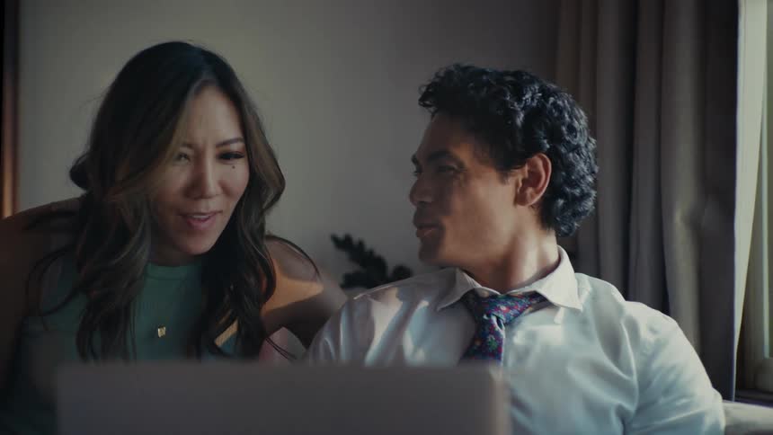 Man sitting interacting with a laptop and a woman coming up beside him