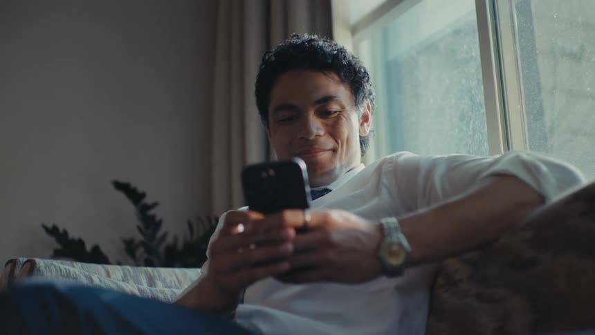 A man sitting on a couch texting on his phone laughing and smiling