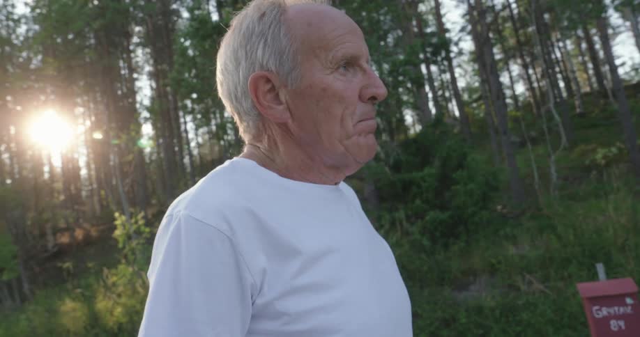 An older man in a white tshirt standing outdoors looking contemplatively