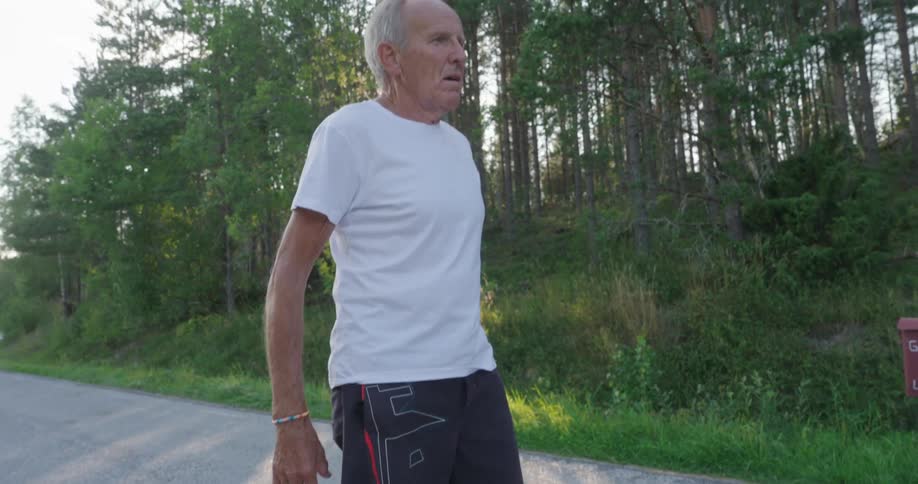 An elderly man in a white shirt is standing on a concrete path with grass on either side