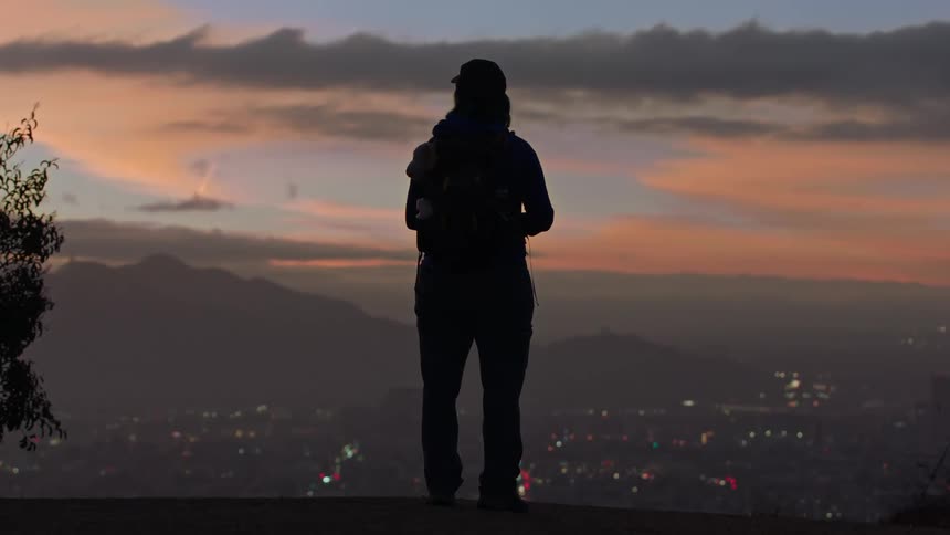 Silhouette of a person standing on the edge of an overlook looking out over a city below as the sun sets