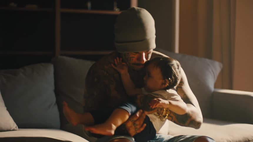 A father with tattoos all over holding their baby on a couch as the child squirms