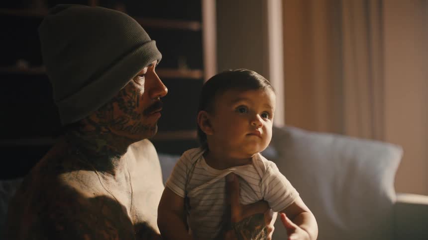 A tattooed man sitting on a couch at home holds a baby in his arms