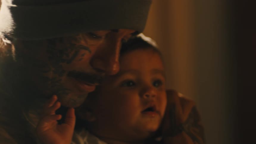 Shirtless tattooed man holds a baby in his arms while seated on a couch in a cozy living room