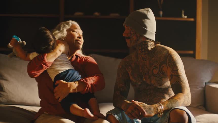 A tattooed man sits on the couch with his mother who holds his grandson in her arms