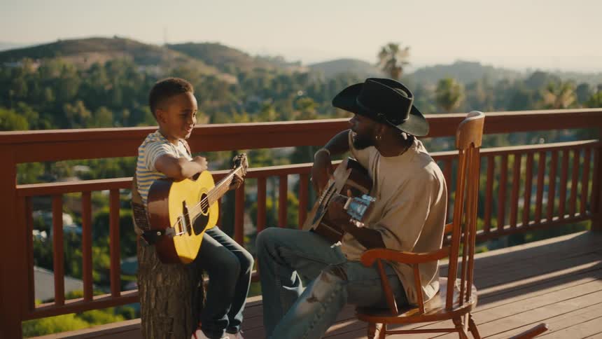 A father and son both holding guitars sitting on a porch talking and playing together
