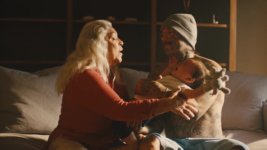 A man in tattoos holding a child before handing it over to a woman who sits on the next to him