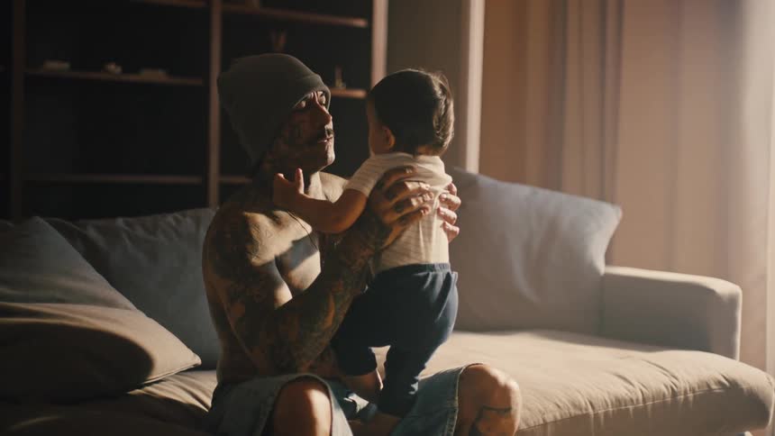 Shirtless tattooed man holds a baby in his arms while seated on a couch in a cozy living room