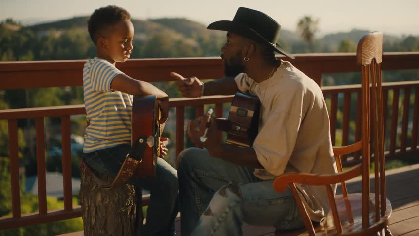 A man sitting on a balcony in the woods teaching his son how to play guitar