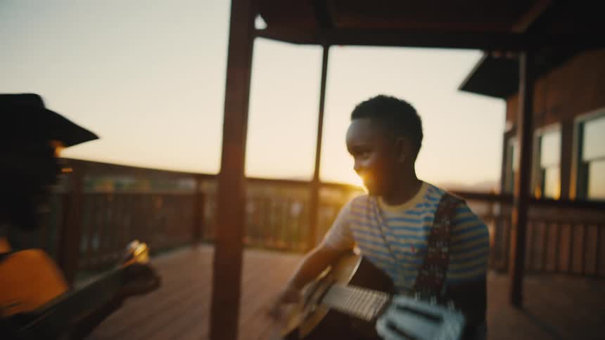 Father and son playing guitar together on a wooden deck at sunset