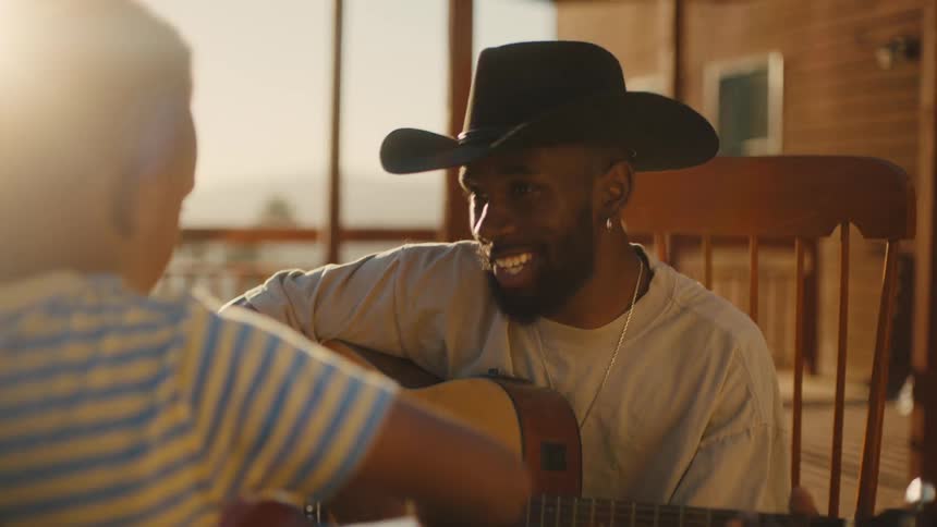 A man in a cowboy hat is playing the guitar while singing happily to a boy in front of him