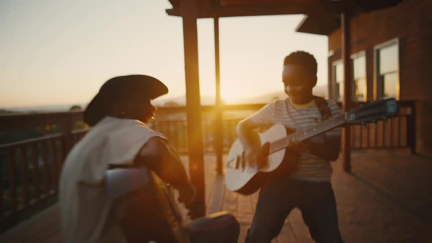 A father and son playing guitar together on a large porch as the sun sets in the distance