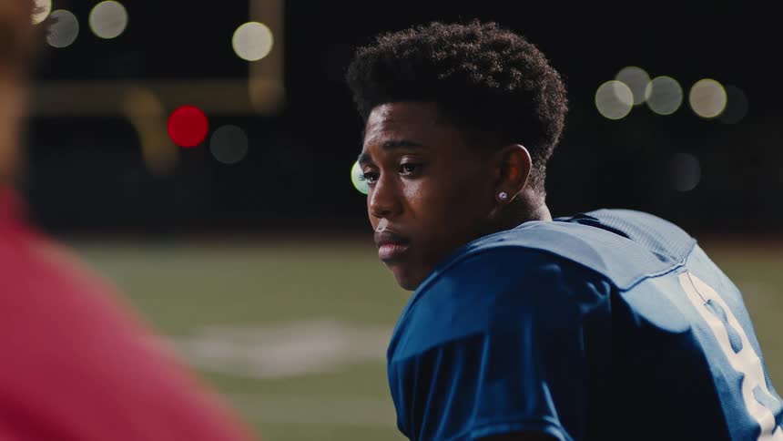 A teenage boy in football sportswear is looking at someone who is talking next to him and has a sad expression on his face