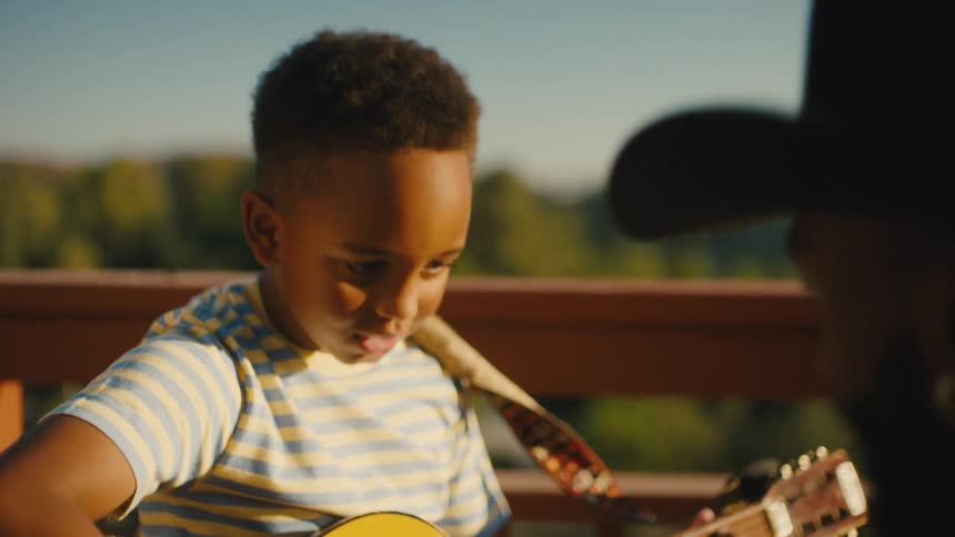 A father teaches his son to play guitar outdoors on a sunny day