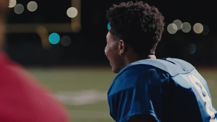 A football player in a blue jersey sitting on the side line smiling and laughing as he throws a football to himself