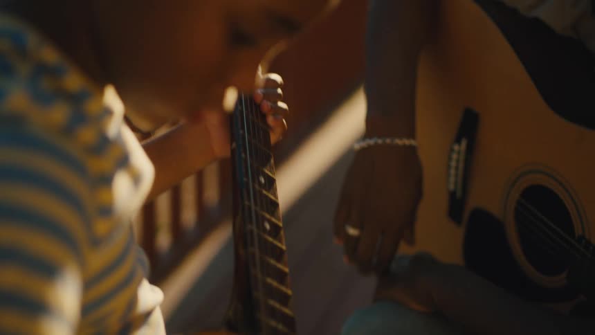 A father teaches his son to play guitar outdoors on a sunny day
