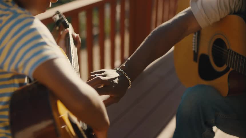 Father sitting on the balcony of his rural home smiles while teaching his son how to play the guitar