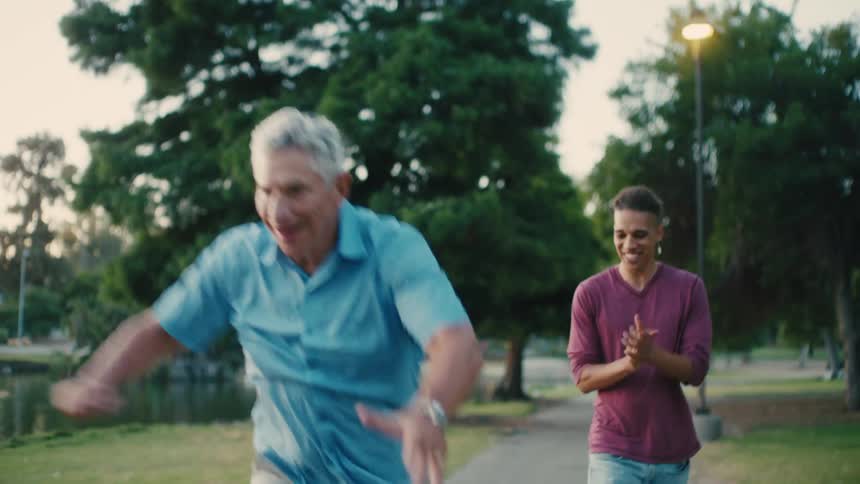 A young man helps an elderly man learn to skateboard at dusk