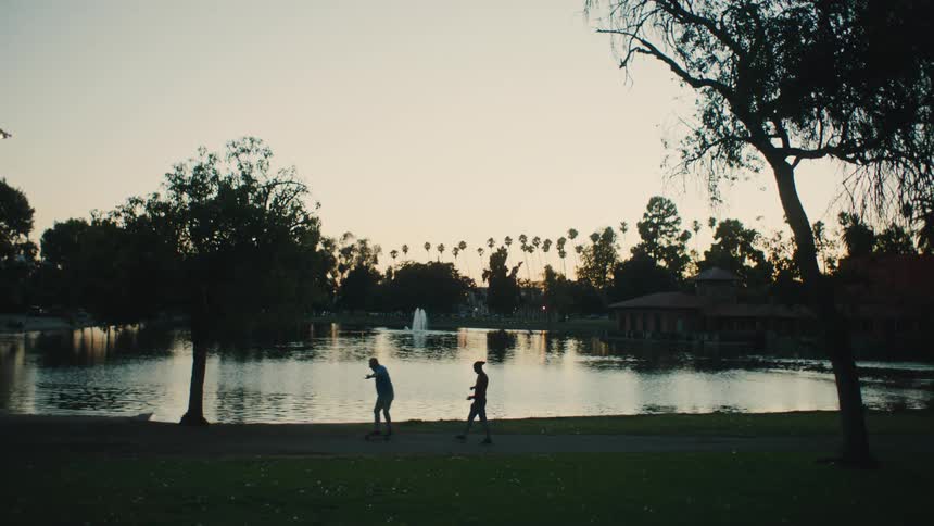 Silhouette of two people in a park skateboarding near a pond at dusk